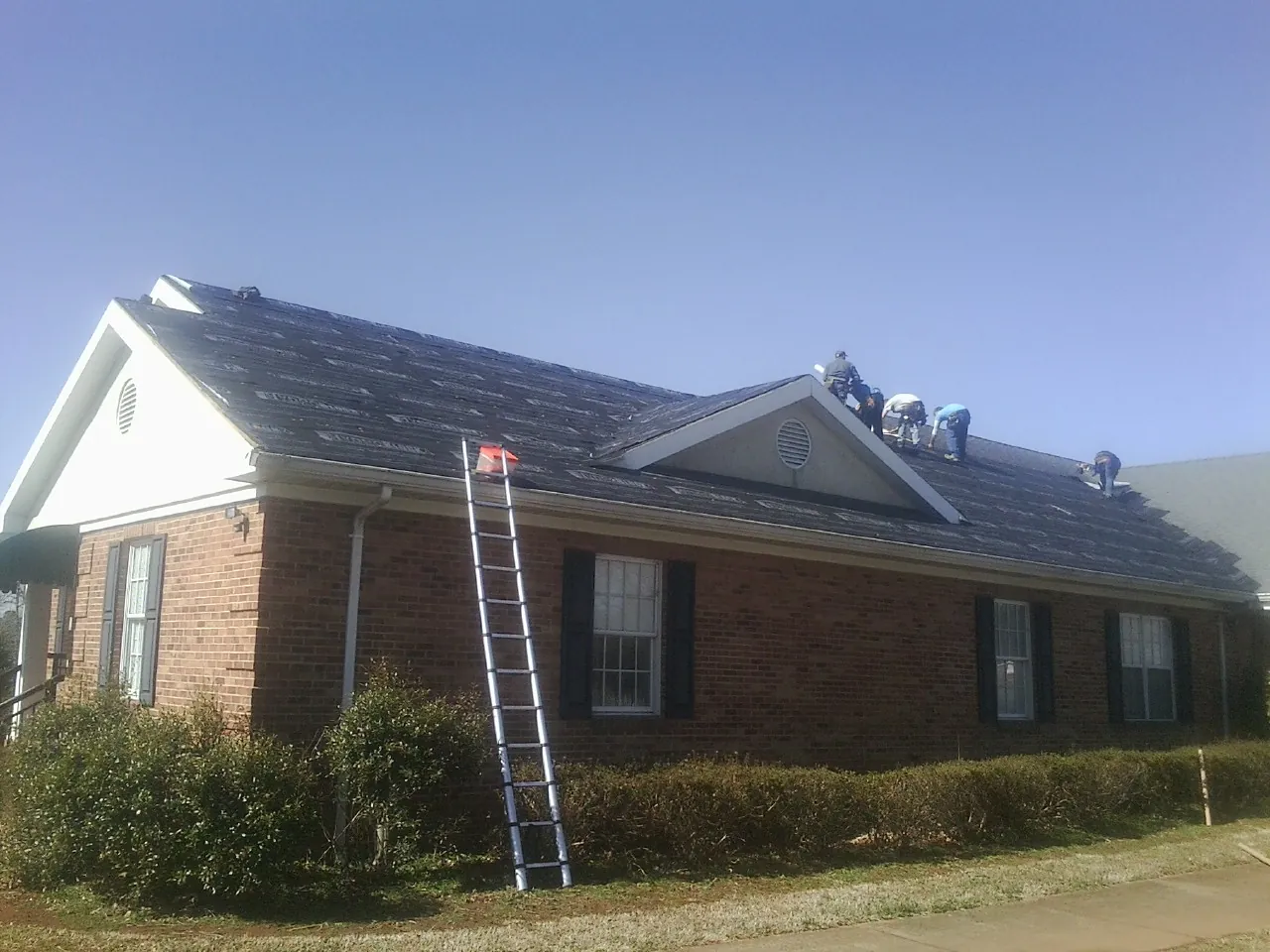 Crew tearing shingles off a brick ranch roof with a ladder set — Metal Roof Repair work by our crew in Fort Plain