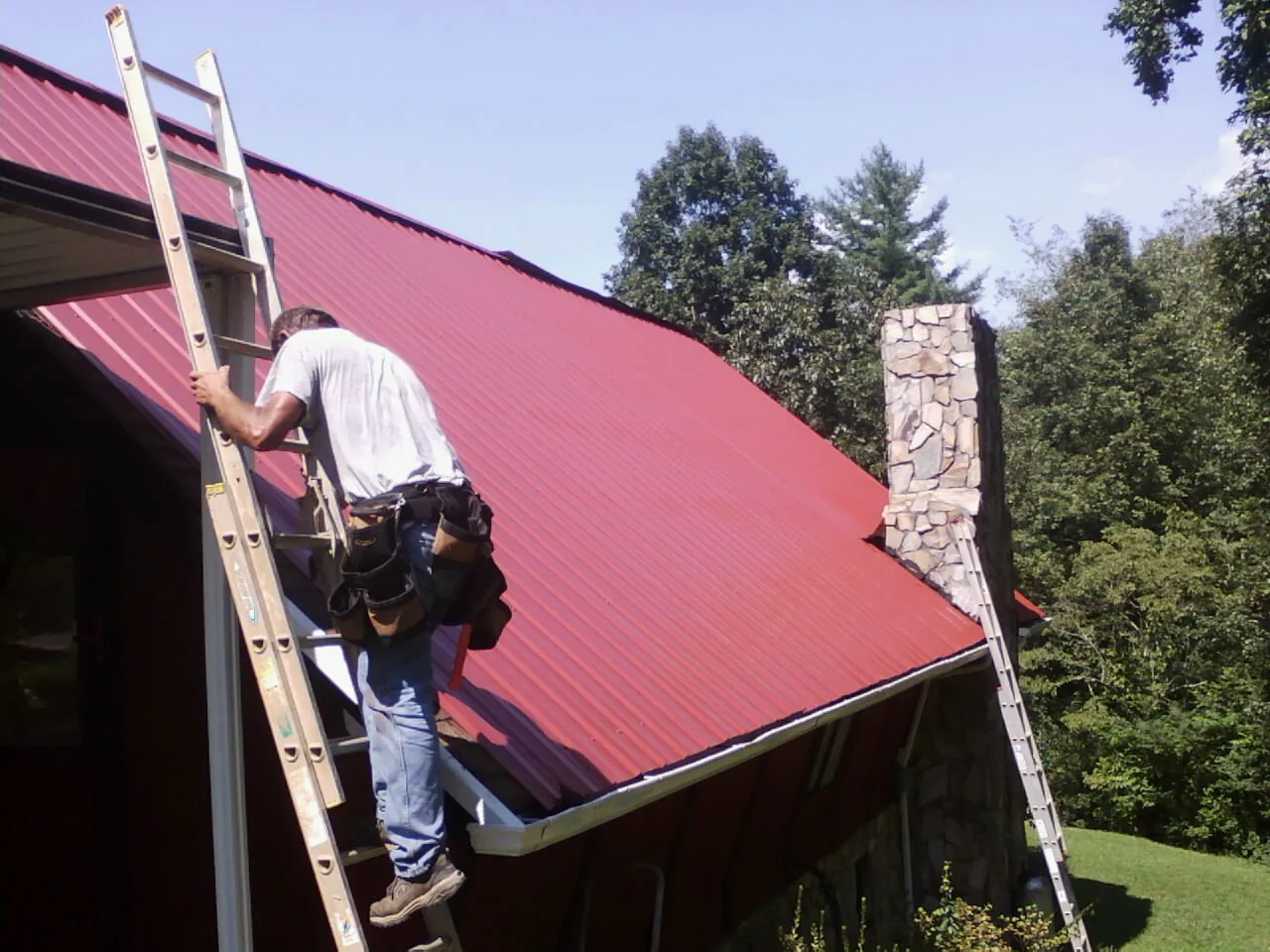 Roofer on a ladder beside a red metal roof and stone chimney — Soffit & Fascia Repair by Roofing Brothers in Fort Plain